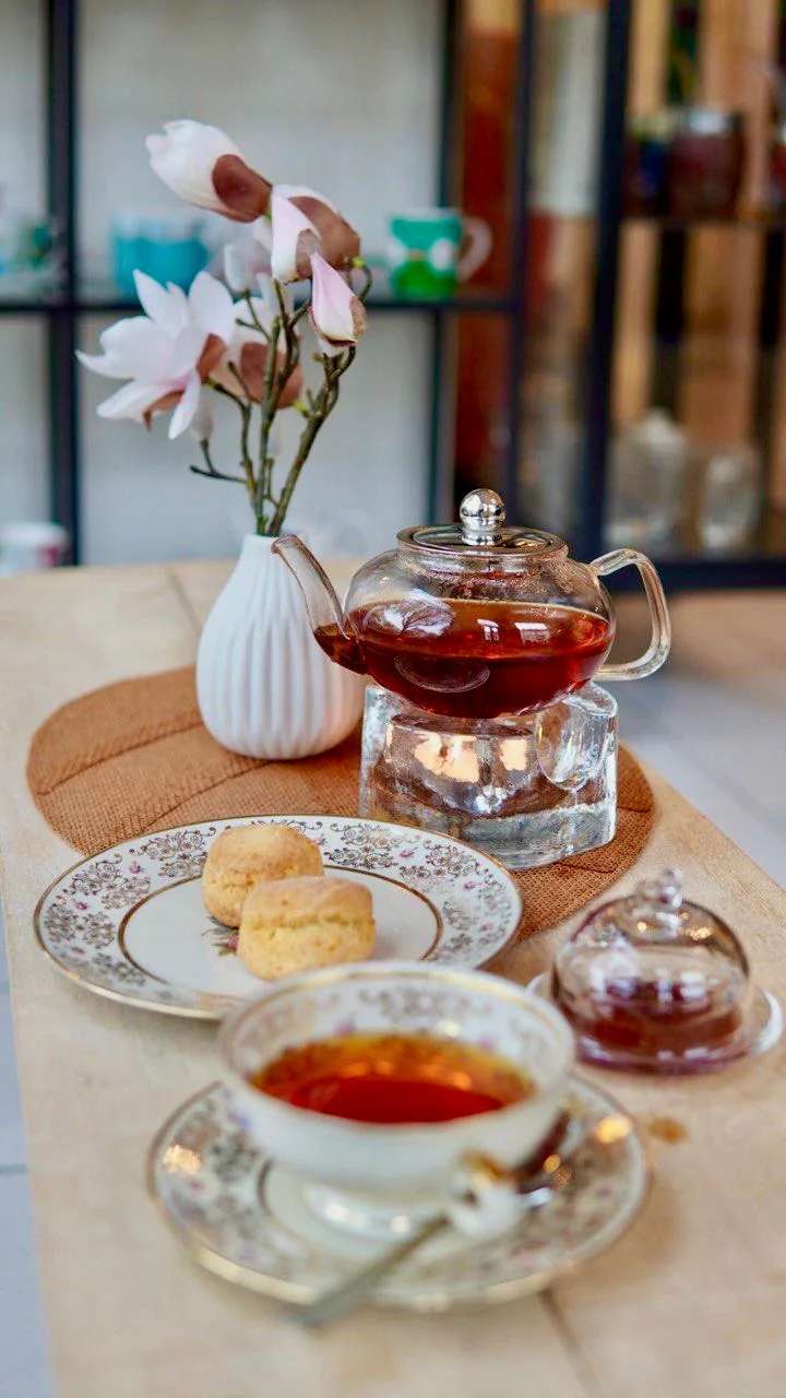 Eine Kanne Tee auf einem Stövchen, florales Porzellan und Scones neben Magnolien